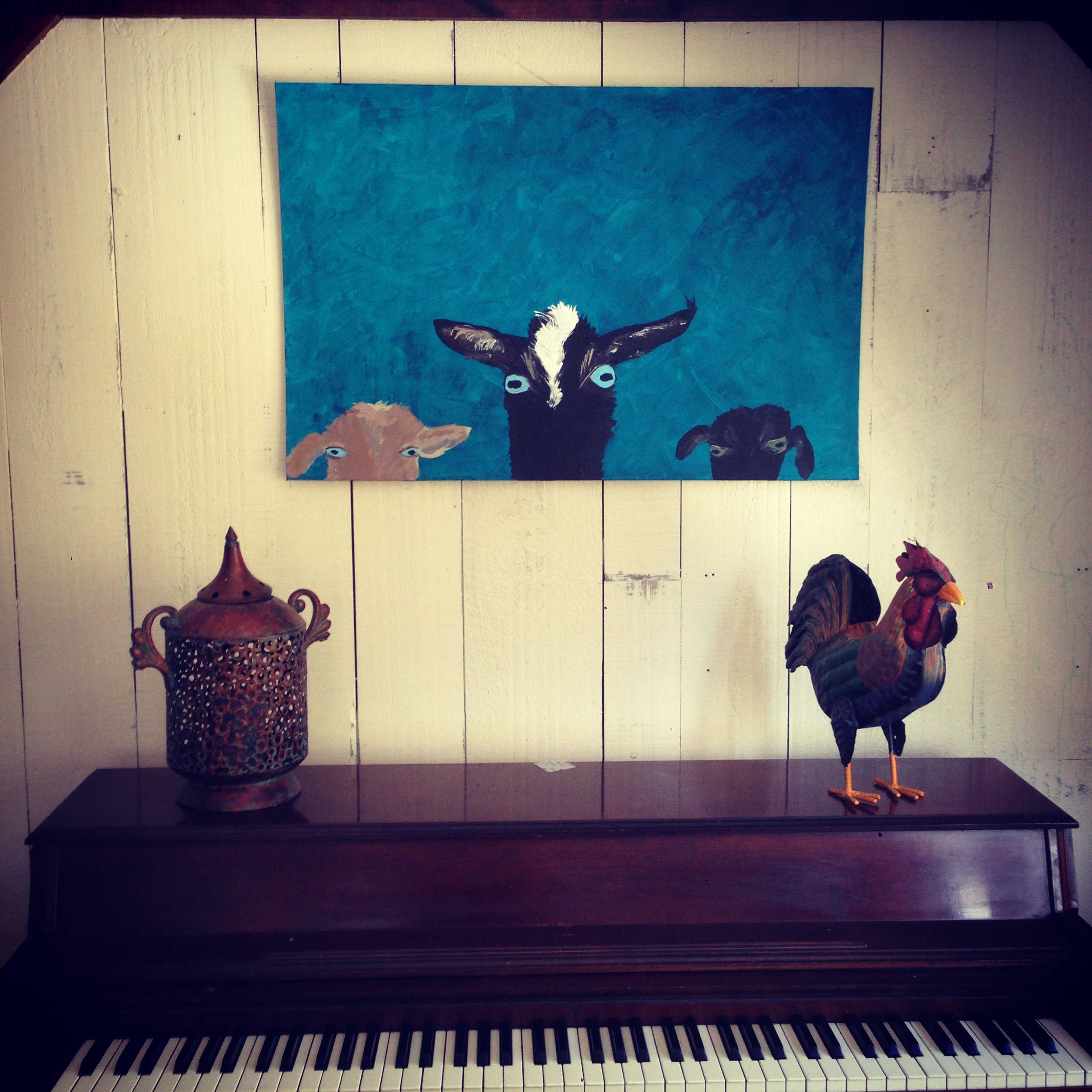 a painting of 3 nigerian dwarf goats against a blue backround hung above a piano